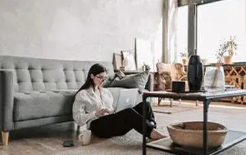 خانمی در حال کار کردن با لپ تاپ که در پذیرایی منزل نشسته و به مبلی با کیفیت تکیه داده است . 