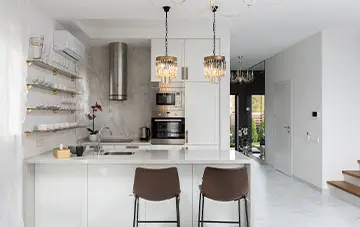آشپزخانه ای که تم آن سفید می باشد و دارای صندلی های قهوه ای و وسایل مشکی میباشد و دو عدد لامپ از سقف آویزان شده است